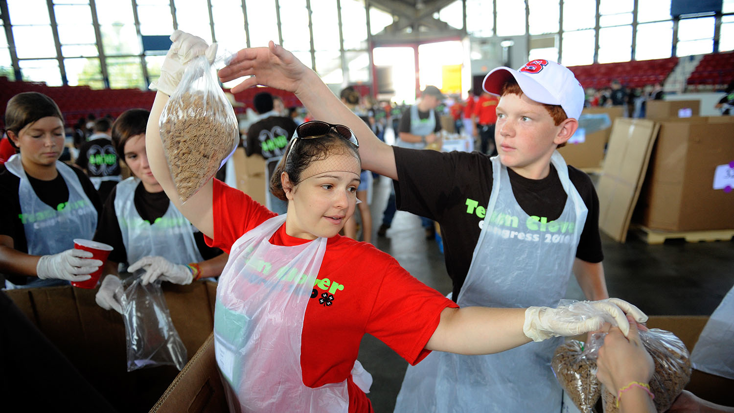 One 4-H student hands a bag of granola to another 4-H student, both wearing aprons.