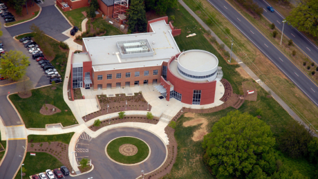 NC State's Carroll Joyner Visitors Center as viewed from above, surrounded by green grass with a circular driveway in front and a diagonal stripe of four-lane highway providing geometric contrast to the upper right.