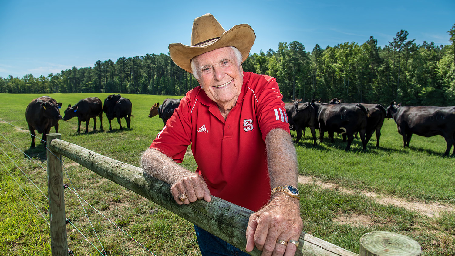 A man named Carroll Joyner wearing a cowboy hat and a red NC State polo shirt smiles at you from behind a cattle fence, green grass and a cloudless blue sky behind him.