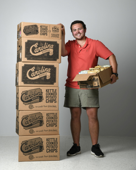 1in6 Snacks founder Josh Monahan stands next to a pile of chips boxes, grinning.