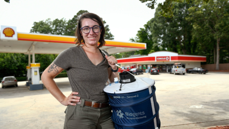 A woman with a cool mosquito tattoo and glasses carries a mosquito trap in front of a gas station.