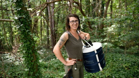 A smiling woman with a mosquito tattoo stands in a forest holding a large mosquito trap.