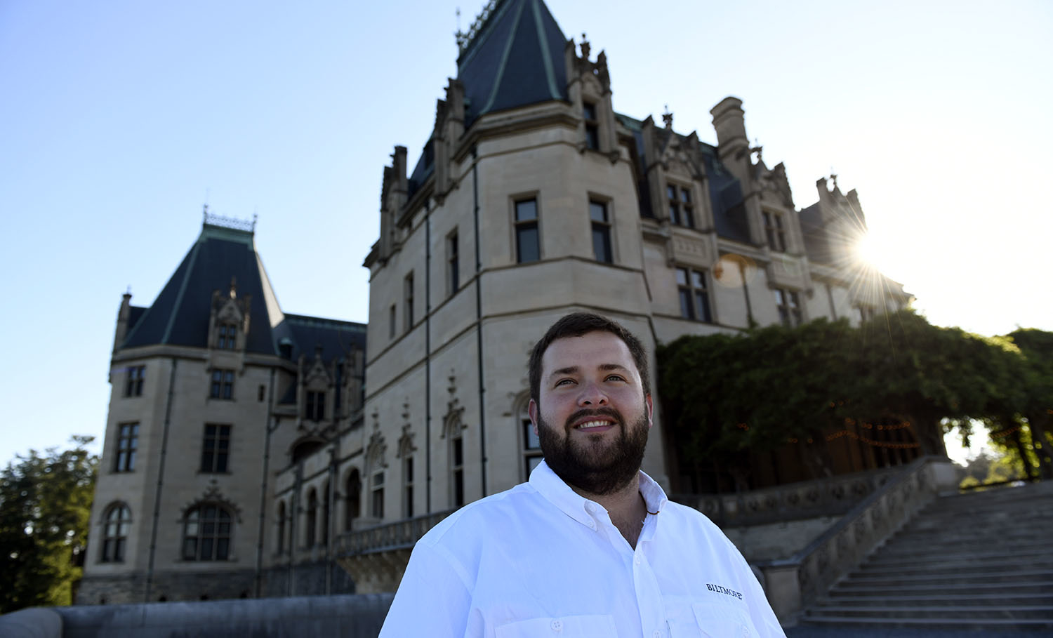 A bearded man stands in the foreground with Biltmore House behind him.